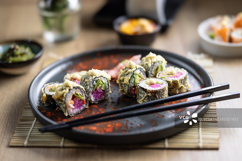 六块炸寿司，也称为天妇罗或热卷，盛放在一个深色、锈色的盘子里。寿司卷外面裹着酥脆的金黄色天妇罗，顶部点缀着一滴日式蛋黄酱。图片素材