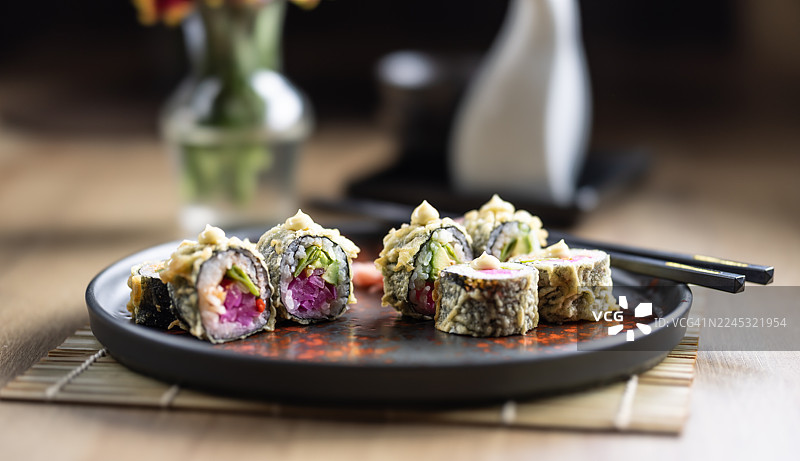 六块炸寿司，也称为天妇罗或热卷，盛放在一个深色、锈色的盘子里。寿司卷外面裹着酥脆的金黄色天妇罗，顶部点缀着一滴日式蛋黄酱。图片素材