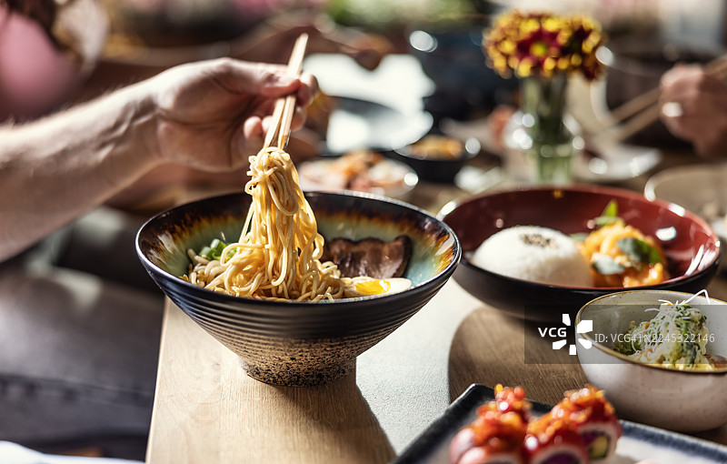 一碗热气腾腾的拉面特写，一只手正用筷子夹起一把面条。前景中模糊的其他菜肴，如寿司和一碗米饭，暗示着在一家亚洲餐厅用餐。图片素材