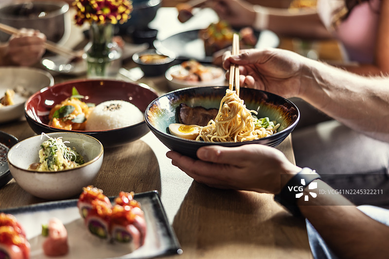 一碗热腾腾的拉面的特写，一个人用筷子夹着面条。前景中模糊的其他菜肴，如寿司和一碗米饭，暗示着在一家亚洲餐厅用餐。图片素材