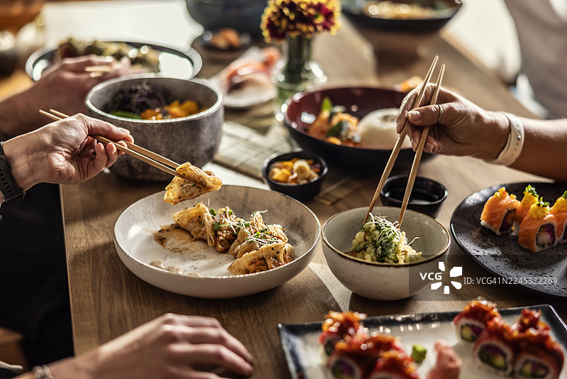 餐桌上摆满了各式亚洲美食，寿司和小碗配菜占据了前景。画面中还有几双拿着筷子的手，暗示着在餐厅里积极的交谈和共享用餐的愉悦。图片素材