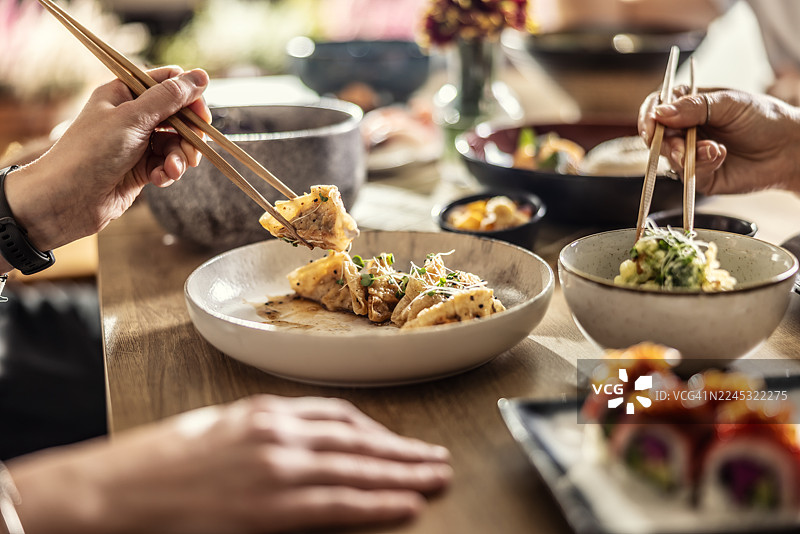 餐桌上摆满了各式亚洲菜肴，寿司和其他小碟配菜占据了前景。画面中有几只拿着筷子的手，暗示着在餐厅里热烈交谈和共享美食的愉悦时光。图片素材