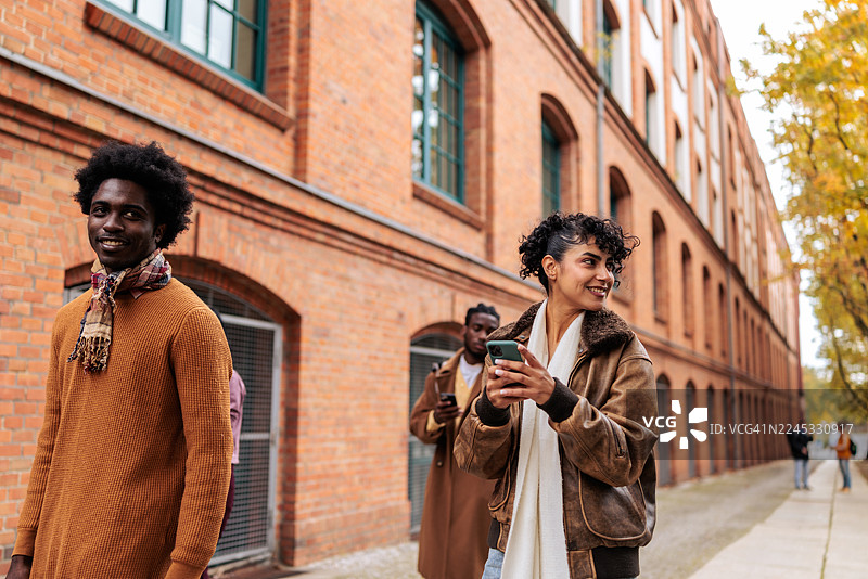 快乐的女人在城市街道上与朋友同行时使用智能手机图片素材