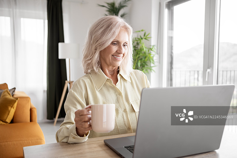 一位微笑的老年女士正在用笔记本电脑喝咖啡和浏览图片素材