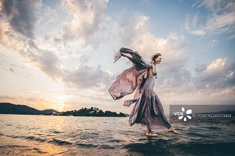 身着飘逸长裙的快乐女子，裙摆在空中飞扬，她优雅地走在浅水中，背景是绚丽的日落。传达了自由、宁静以及人与自然元素的美好互动。图片素材