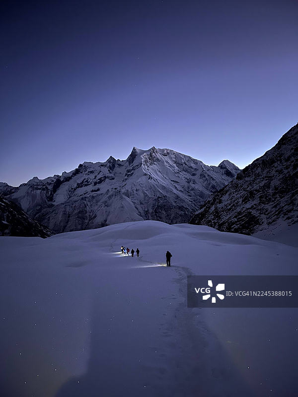 徒步者头戴矿灯，在黎明时分穿越雪山小径，背景是雄伟的雪峰。图片素材