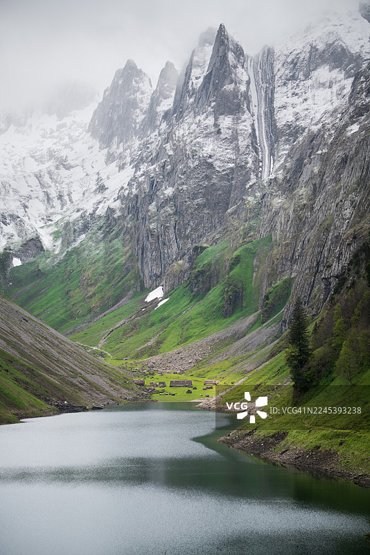 瑞士戏剧性的山地景观，宁静的湖面倒映着天空，周围环绕着郁郁葱葱的绿色山谷和白雪皑皑的山峰，天空多云。图片素材