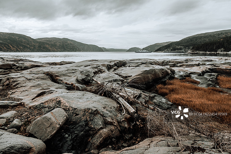 阴沉天空下，拥有崎岖岩石和平静水面的戏剧性海岸风光，远处有一艘渡轮。图片素材