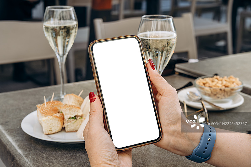 女性在咖啡馆手持带有空白屏幕的手机模型，周围摆放着精致的饮品和美味的开胃菜，非常适合设计使用图片素材