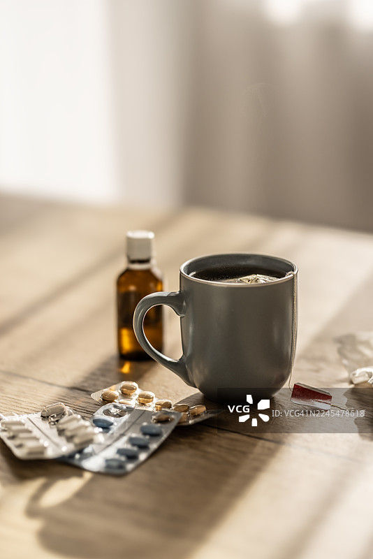 厨房的桌子上放着一个冒着热气的茶包，表明它还很烫。杯子旁边是药片泡罩包装、一瓶药水和一个揉皱的手帕。图片素材