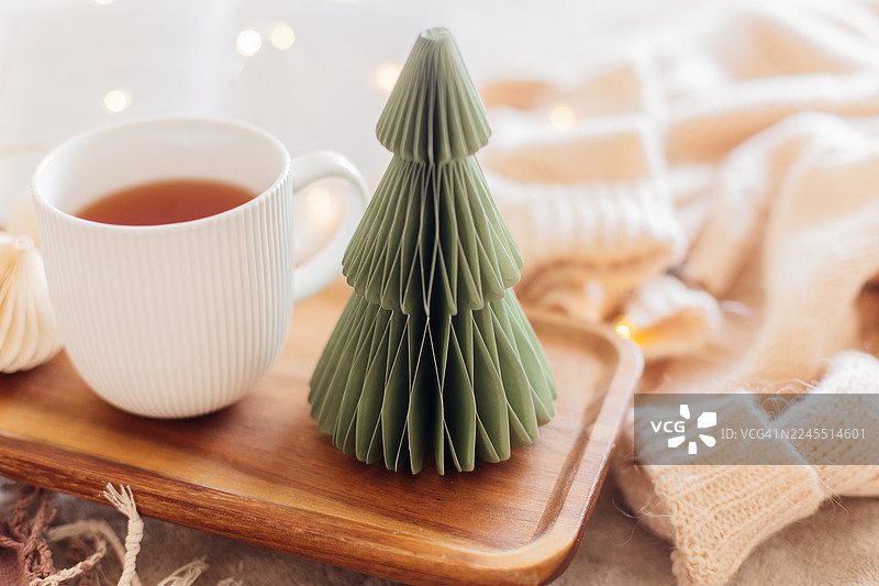 一个绿色的纸质圣诞树装饰品放在木质托盘上，旁边放着一个白色的茶杯，周围是舒适的针织物品，营造出温馨诱人的节日氛围图片素材