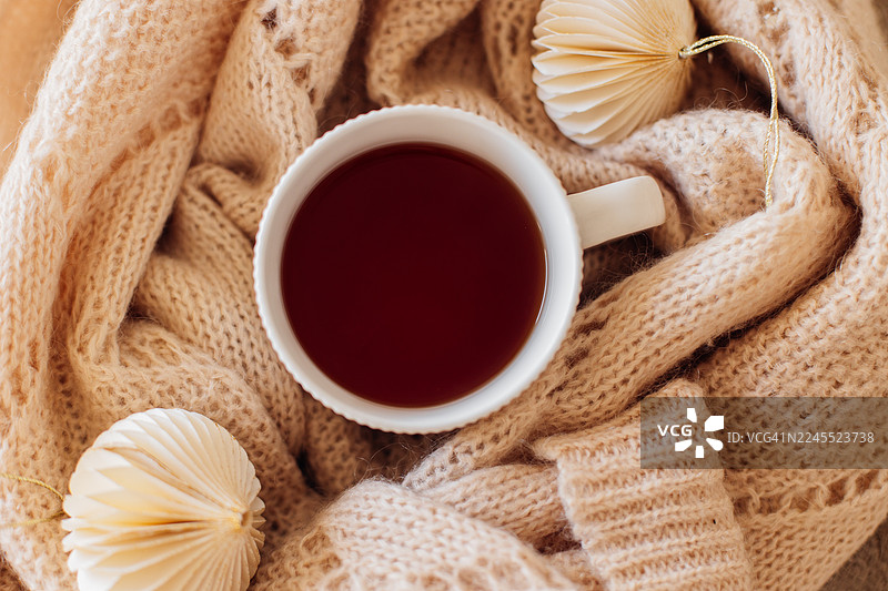 温暖的茶杯放在舒适的针织围巾上，周围装饰着纸质摆件，营造出宁静的氛围，非常适合冬季放松和舒适。图片素材