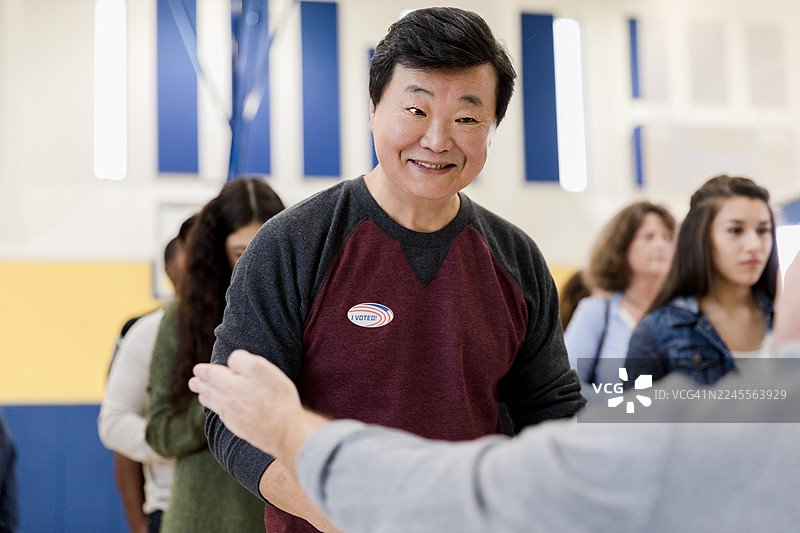 微笑着的男人自豪地戴着“我已投票”贴纸排队，体现了公民参与和投票精神。图片素材