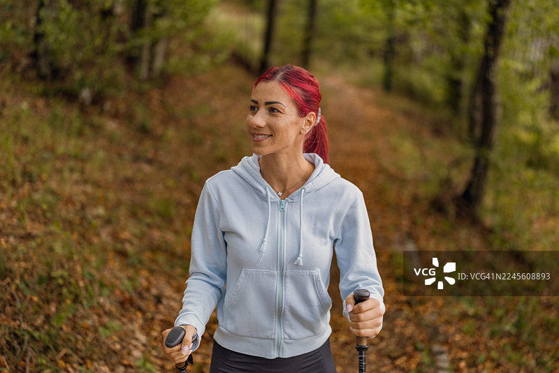 粉色头发的运动女孩在森林里的道路上使用登山杖徒步。图片素材