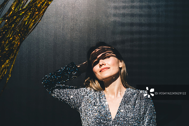 身穿银色亮片连衣裙的微笑女性肖像，背景为深色，在温暖阳光的照射下，泛着柔和的闪光和阴影。图片素材