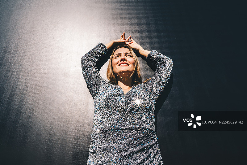 身穿银色亮片裙的微笑女性肖像，站在深色背景前，沐浴在温暖的阳光下，营造出柔和的闪光和阴影。图片素材