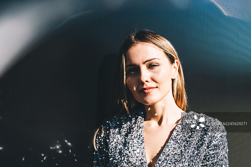 一位身穿银色亮片连衣裙的微笑女性肖像，站在深色背景前，温暖的阳光照亮她，营造出柔和的闪光和阴影。图片素材