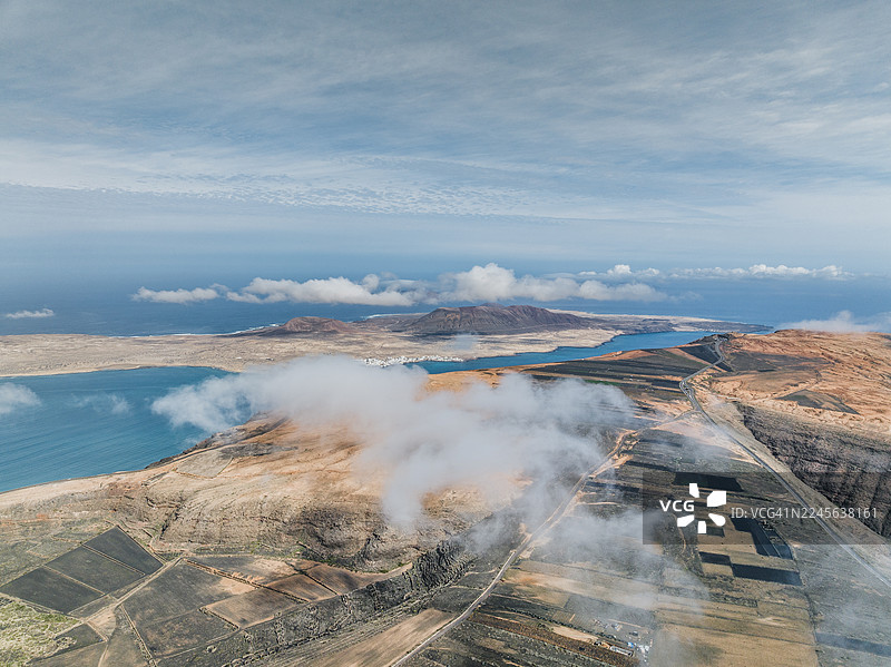 航拍图，展示了低空云层漂浮在悬崖顶部的农田上，背景是西班牙加那利群岛兰萨罗特岛的格拉西奥萨岛。图片素材