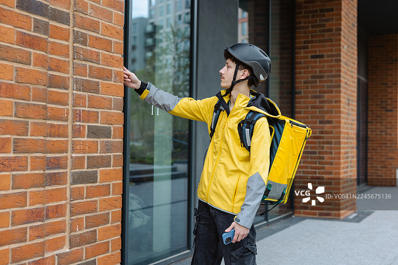 穿着亮黄色夹克的送餐骑手在砖砌建筑外按响了楼宇对讲门铃图片素材