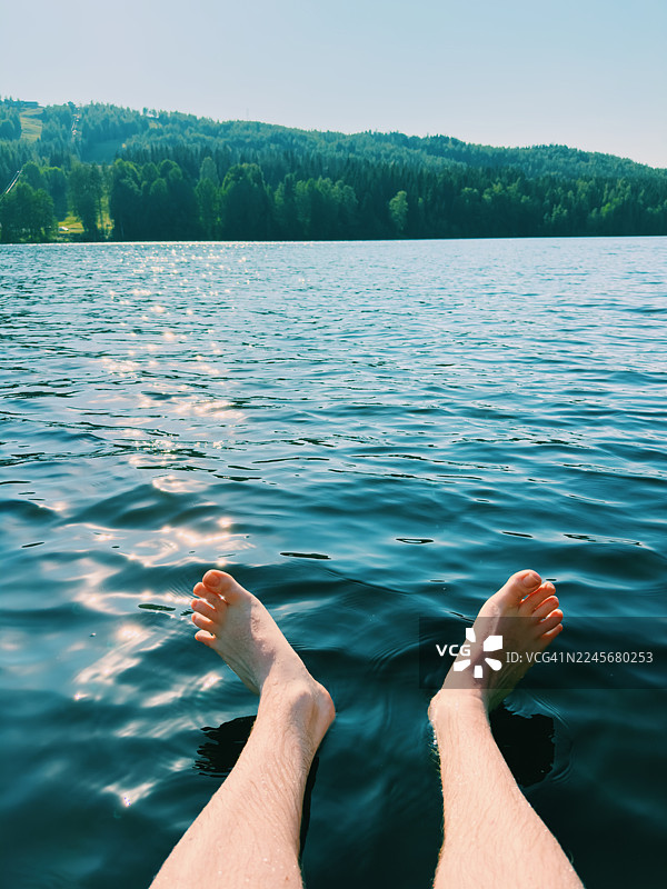 从第一人称视角拍摄的画面，展示了在芬兰北部阳光明媚的夏日里，男人的双脚浸入湖水中。图片素材
