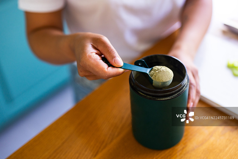 厨房台面上，一个人正在往绿色摇摇杯里舀蛋白粉，为健身做准备。图片素材