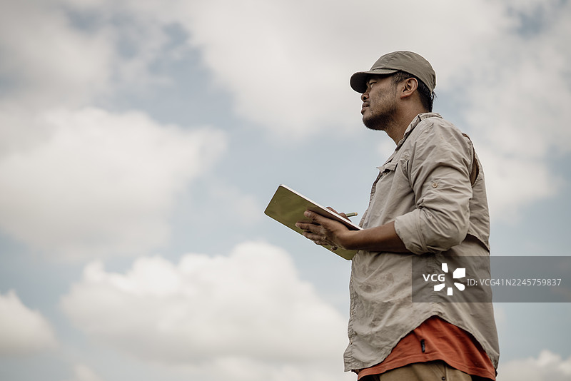 一位农民或农业研究员跪在绿色的稻田中，在明亮的蓝天下观察作物并用平板电脑记录数据。图片素材