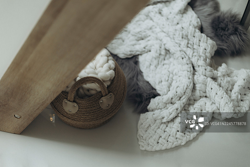 米白色粗针织盖毯和编织篮子放在灰色人造毛皮地毯旁边浅色的地板上。图片素材