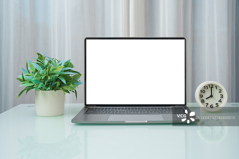 家中的办公桌上放着一台笔记本电脑。极简风格的办公室里，一张木质办公桌上放着一台白色空白屏幕的笔记本电脑、一个闹钟和一盆盆栽，并配有装饰品。图片素材