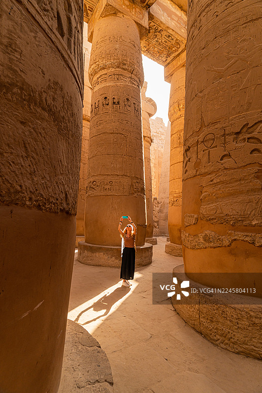 一位女性游客走在卡纳克神庙宏伟柱厅的柱子之间，并用智能手机拍照。图片素材