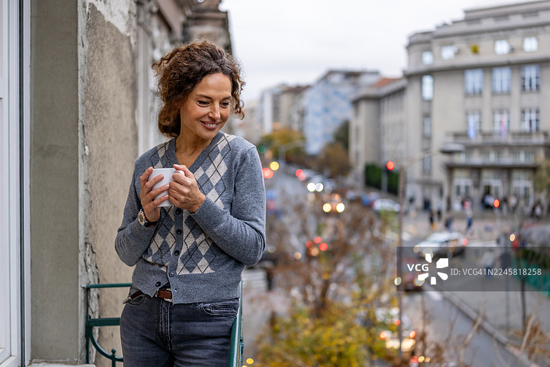秋日傍晚，女子在阳台上悠闲地喝着咖啡，俯瞰着城市街道图片素材