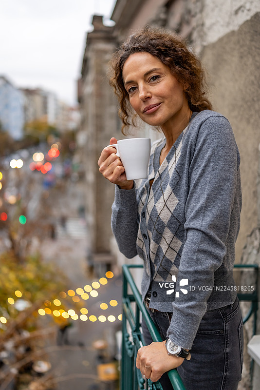 身穿灰色菱格纹开衫的女子在城市阳台上，俯瞰繁忙的街道，悠闲地享用咖啡。图片素材