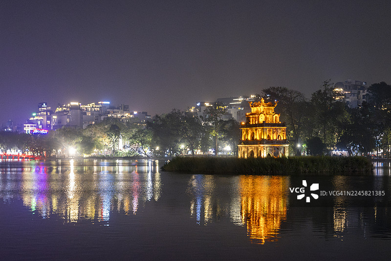 夜色中，还剑湖倒映着灯火辉煌的龟塔和周围的城市建筑，构成了一幅宁静而充满活力的城市景象，位于越南河内。图片素材