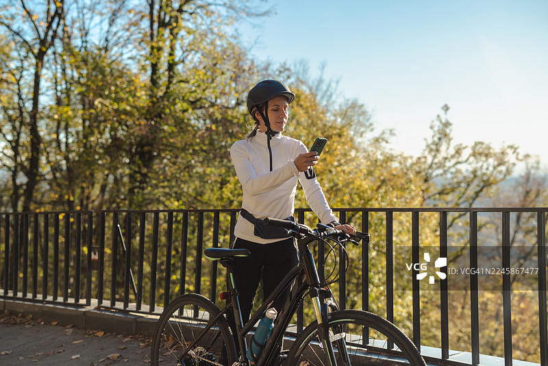 秋季公园里，戴着头盔的女士骑着自行车，同时使用智能手机。图片素材