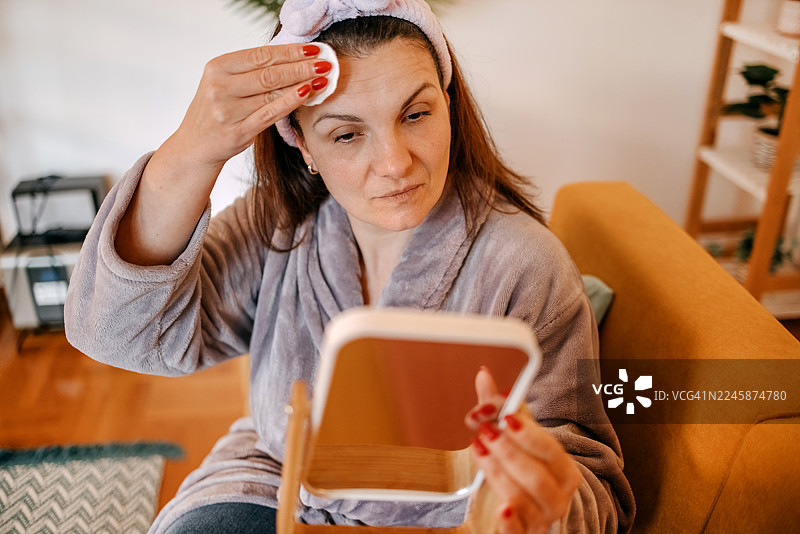 女人坐在客厅沙发上，在镜子前使用面霜清洁面部皮肤，呵护肌肤。图片素材