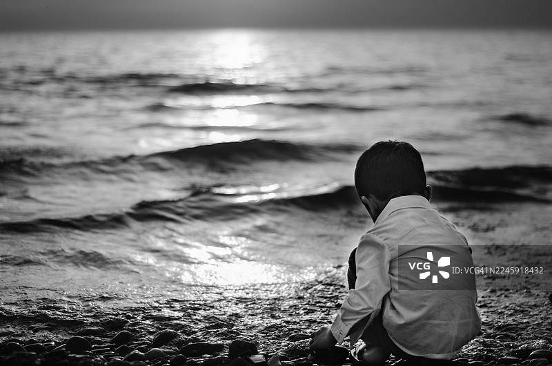 黄昏时分，孩童在岩石海滩上，夕阳的倒影在水面闪耀。低角度拍摄捕捉了轻柔的海浪以及男孩与周围环境的安静互动，营造出宁静与自然之美。图片素材