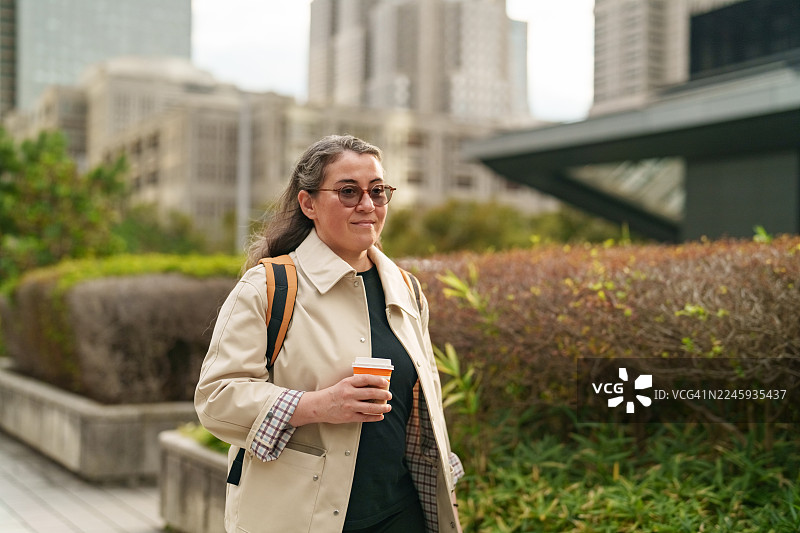 一位女士在现代都市环境中悠闲地散步，手中拿着一杯咖啡。图片素材