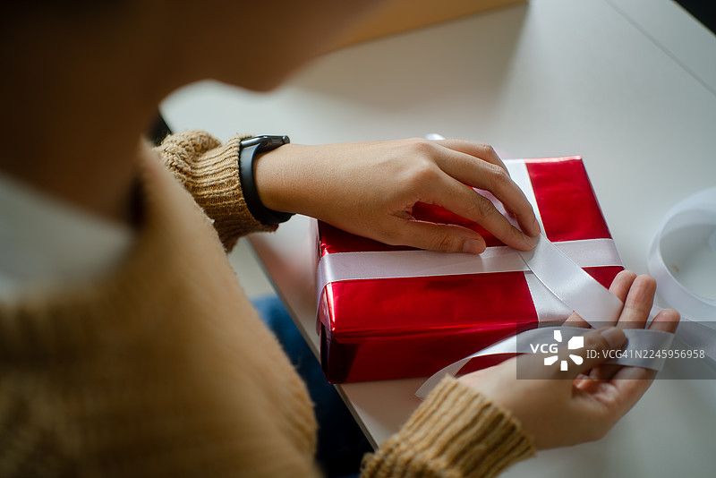 从后面看，一位女士正在为即将到来的年度节日生日惊喜系丝带并包装礼品盒，一位快乐的女士在家里的客厅准备礼物。图片素材