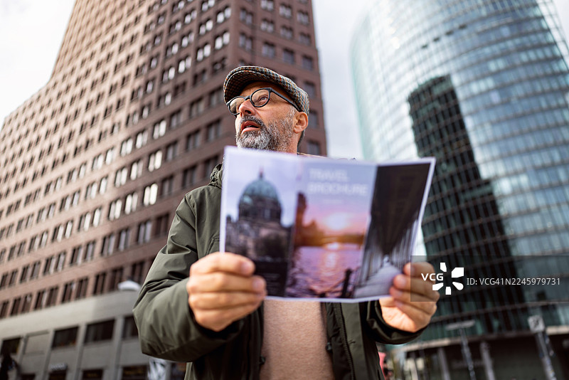 戴眼镜、穿着保暖衣物的中年男性游客在柏林波茨坦广场，手持地图，背景是现代建筑图片素材