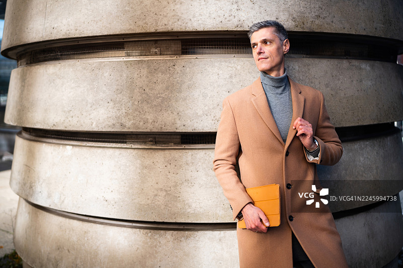 一位专业男士身穿时尚外套，手持平板电脑，倚靠在柏林的一面现代墙壁旁图片素材