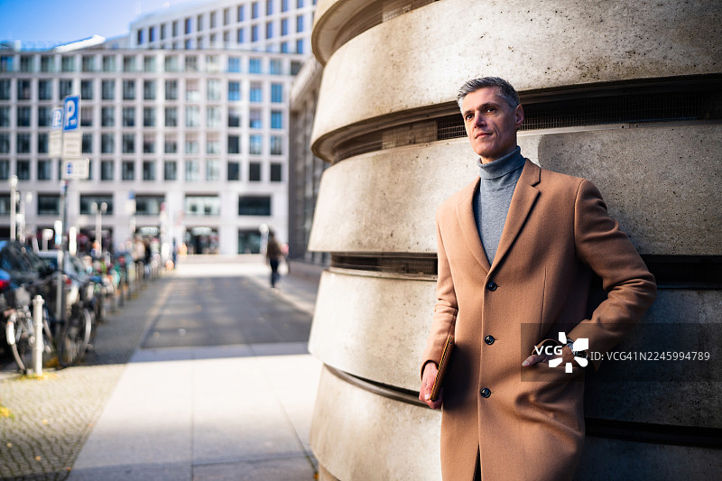一位穿着时尚外套的成熟商人倚靠在柱子旁，一手拿着笔记本电脑，另一只手插在口袋里，背景是柏林波茨坦广场。图片素材