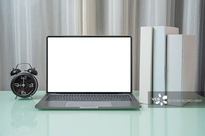 在家里的办公桌上放着一台笔记本电脑。在极简风格的办公室里，木质工作台上放着一台白色空白屏幕的笔记本电脑、一个闹钟和一本书，房间里还有一些装饰品。图片素材
