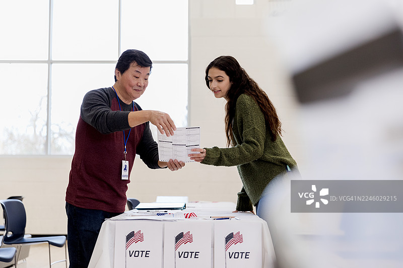 室内投票站，成年志愿者在窗户背景下协助年轻女子投票。图片素材