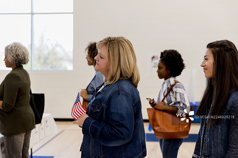一群多元化的女性排队等候，她们手中拿着一面美国国旗和一部手机。图片素材