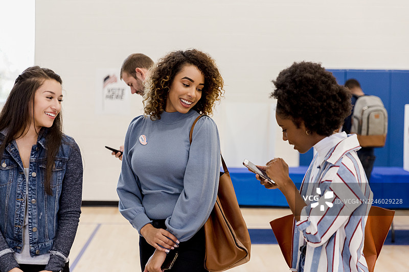 一群来自不同背景的女性在室内随意地聊天，她们看着手机，可以看到她们的投票贴纸。图片素材