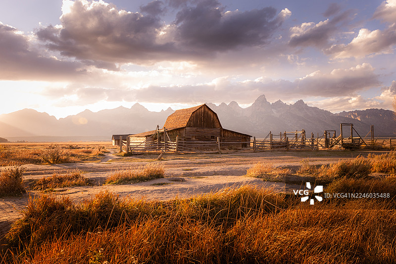 日落时分，金黄色的田野里坐落着乡村谷仓和栅栏，远方是连绵的山峦，位于美国杰克逊大提顿国家公园。图片素材