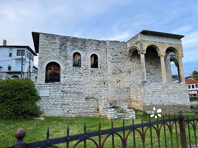 低角度拍摄，一座带有拱形窗户和柱子的古老石头建筑，背景是多云的天空，前景是绿草和装饰性围栏，阿尔巴尼亚图片素材