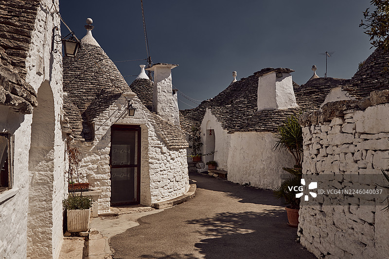 意大利阿尔贝罗贝洛，晴朗蓝天下，狭窄街道旁排列着带圆锥形屋顶的迷人特鲁利小屋图片素材