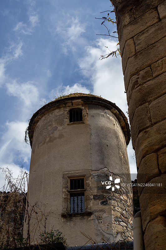 从低角度仰望一座古老、饱经风霜的圆柱形建筑，窗户狭小并带有栏杆，背景是多云的蓝天，另一座石头建筑构成了前景，法国，康塔尔图片素材