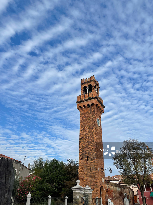 从低角度仰视历史悠久的钟楼，背景是多云的蓝天，周围环绕着传统建筑和茂密的树木，意大利圣皮耶特罗卡里亚诺图片素材