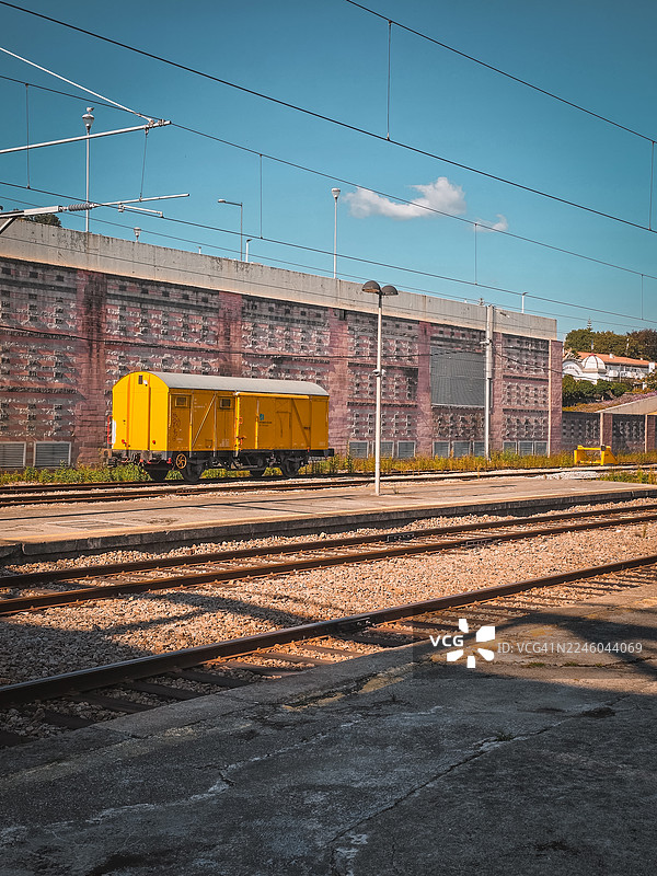 一节鲜黄色的火车车厢静止地停在铁轨上，背景是蓝天、一堵混凝土墙和架空电线。图片素材